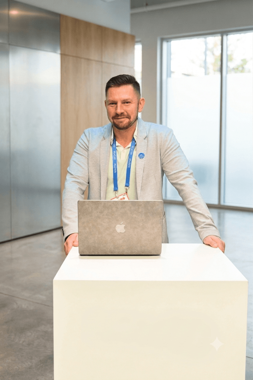 Man in grey blazer standing behind a laptop on a white pedestal in an office.